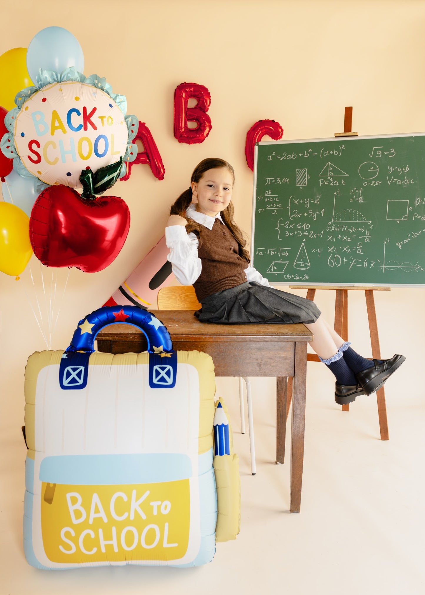 A young girl in a school uniform sits on a wooden desk in a classroom setting, with a large inflatable 'Back to School' backpack in the foreground, red and yellow 'Back to School' balloons, and a chalkboard in the background.