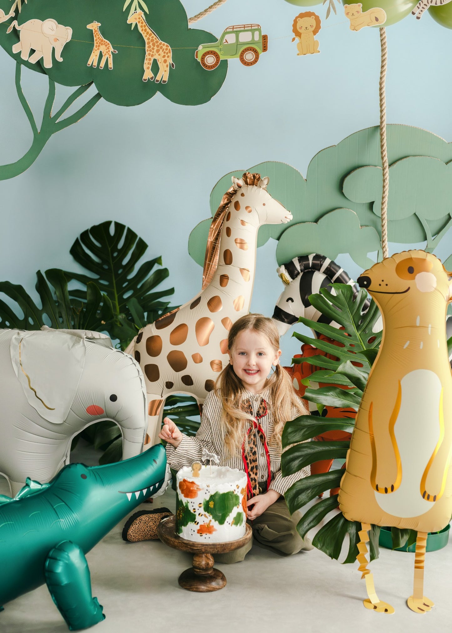 Child with a cake surrounded by inflatable animal decorations in a jungle-themed room.