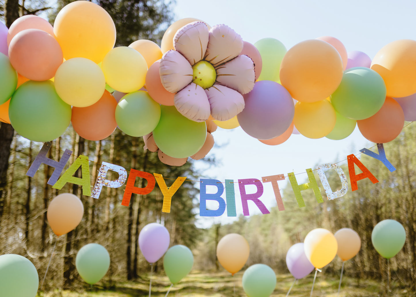 Outdoor birthday party decoration with pastel balloons, large floral balloon, and colorful Happy Birthday banner in the forest