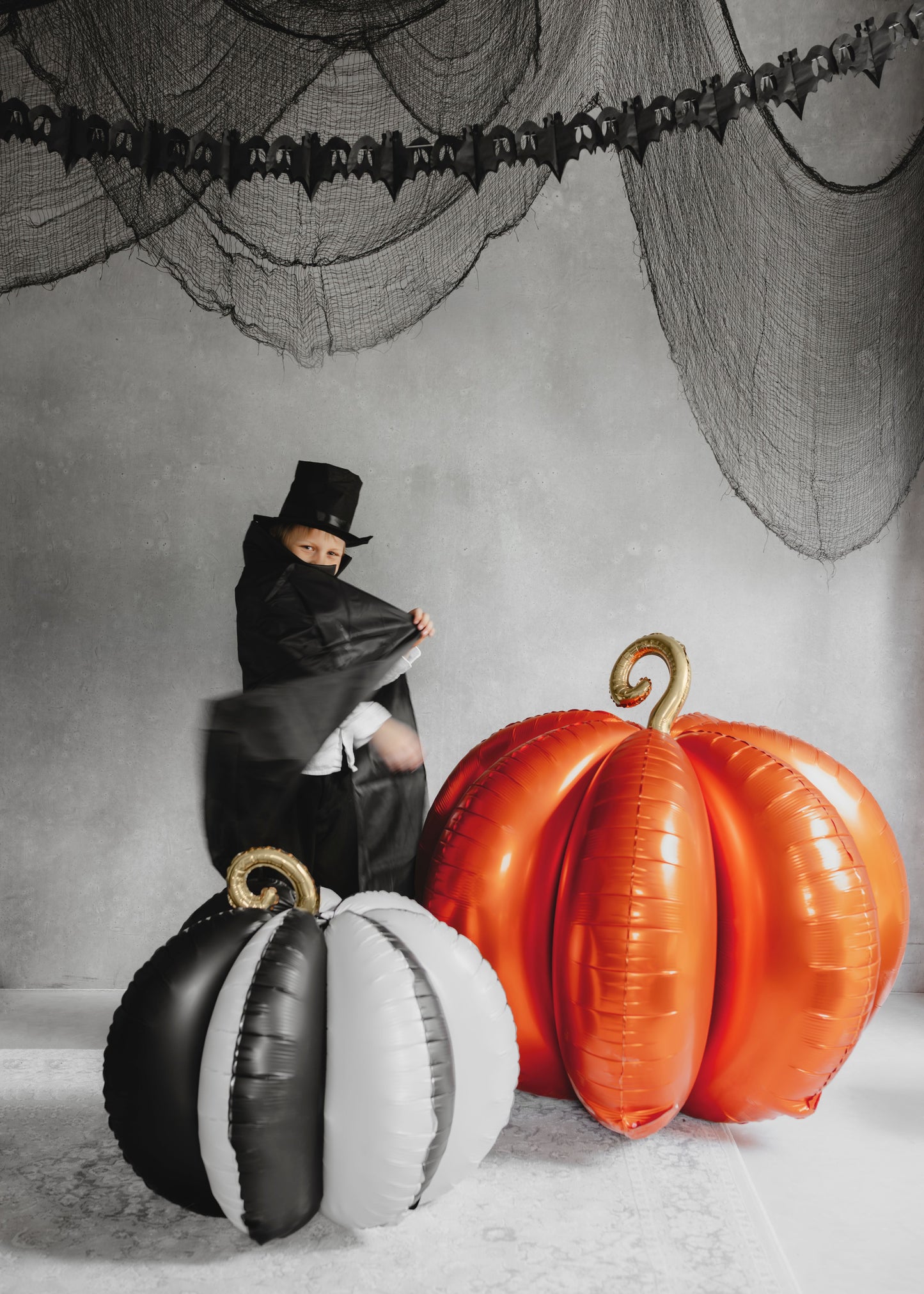 Person in a black outfit with a top hat next to inflatable pumpkins on a Halloween-themed background.