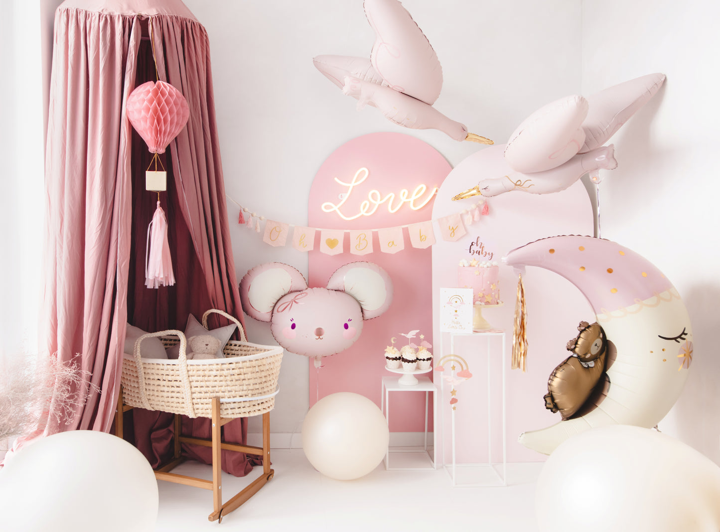 Baby girl welcome party setup in soft pink tones with stork and moon foil balloons, Oh Baby banner, mouse balloon, cake display, and bassinet under canopy – perfect for a newborn celebration.