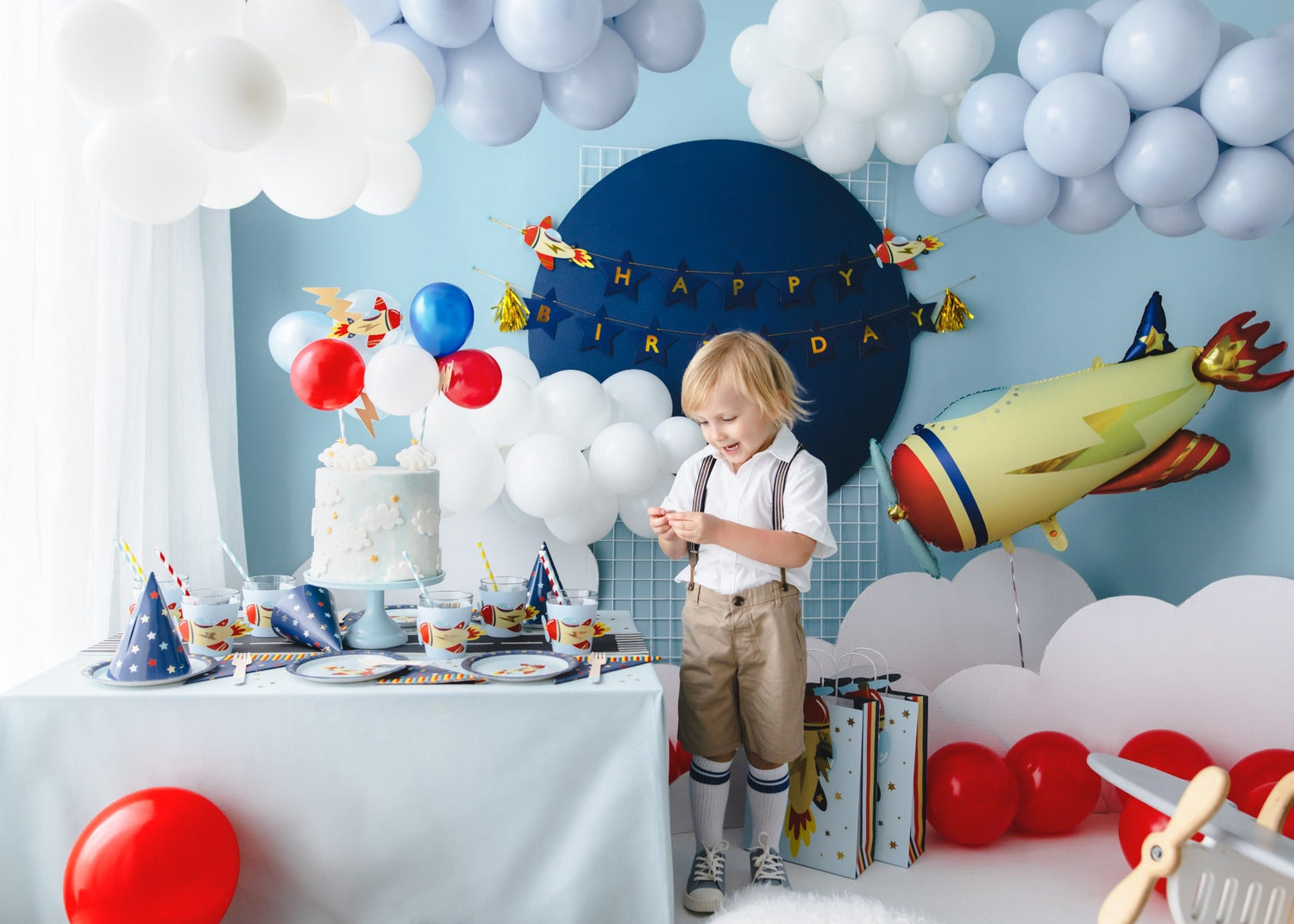 Smiling boy standing at a rocket-themed birthday party with white balloon clouds, colorful decorations, and a large rocket foil balloon
