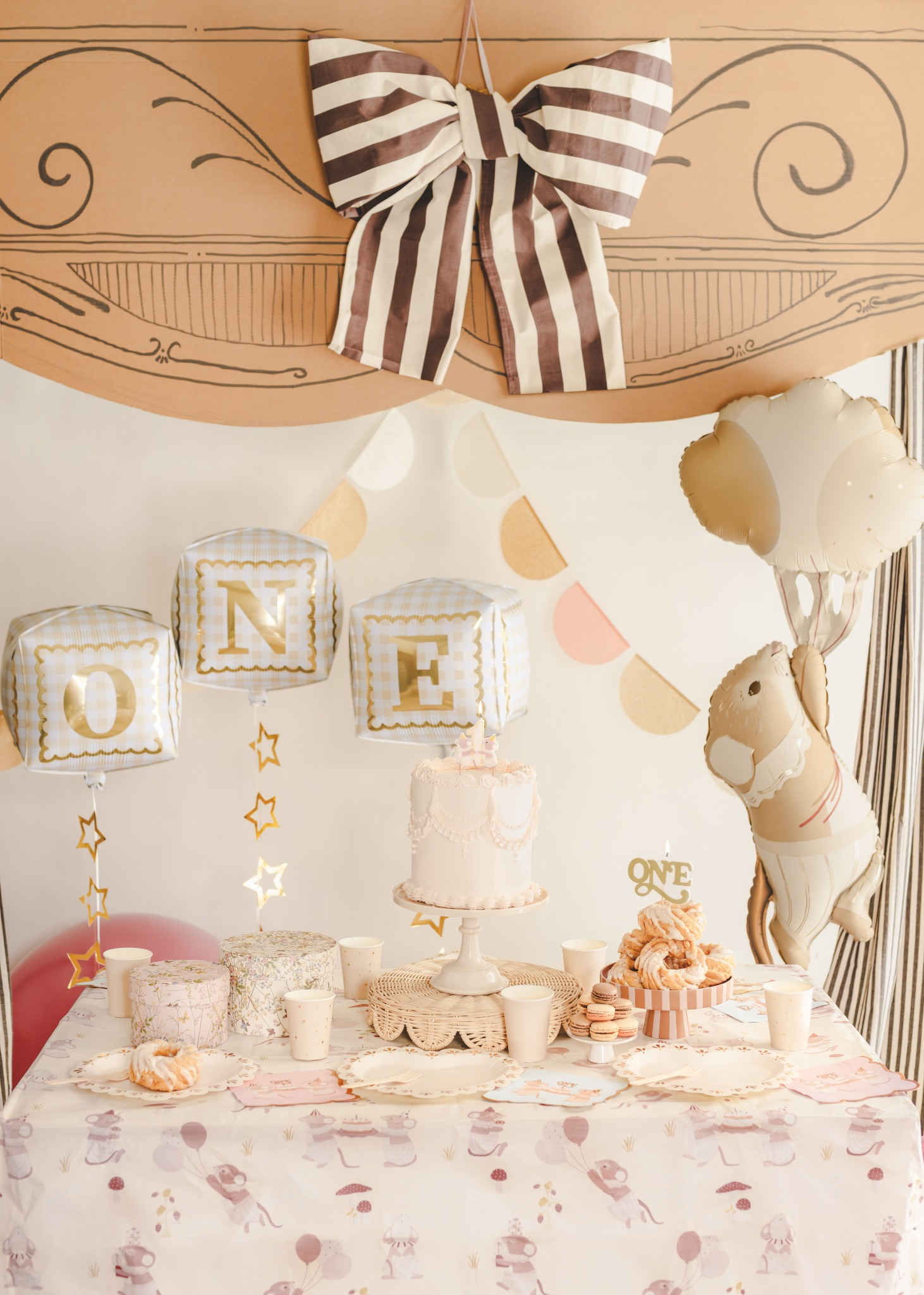 Teddy bear first birthday party decorations with pastel and gold accents, featuring a 'First Birthday' banner, teddy bear balloons, gold star balloons, themed gift bags and a decorated table setup