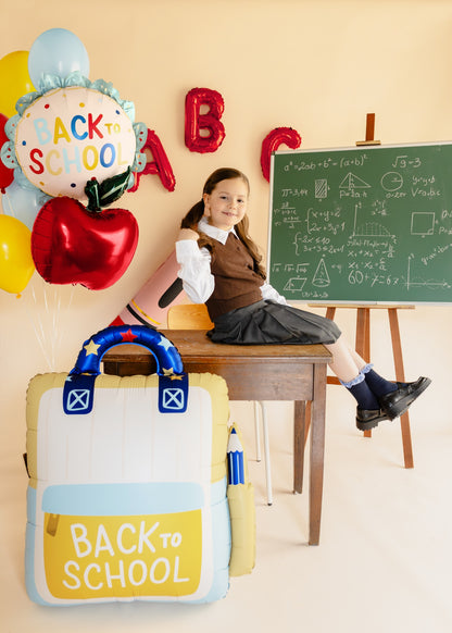 A young girl in a school uniform sits on a wooden desk in a classroom setting, with a large inflatable 'Back to School' backpack in the foreground, red and yellow 'Back to School' balloons, and a chalkboard in the background.