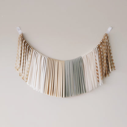 Decorative fabric banner with fringe in neutral colors hanging against a plain wall.