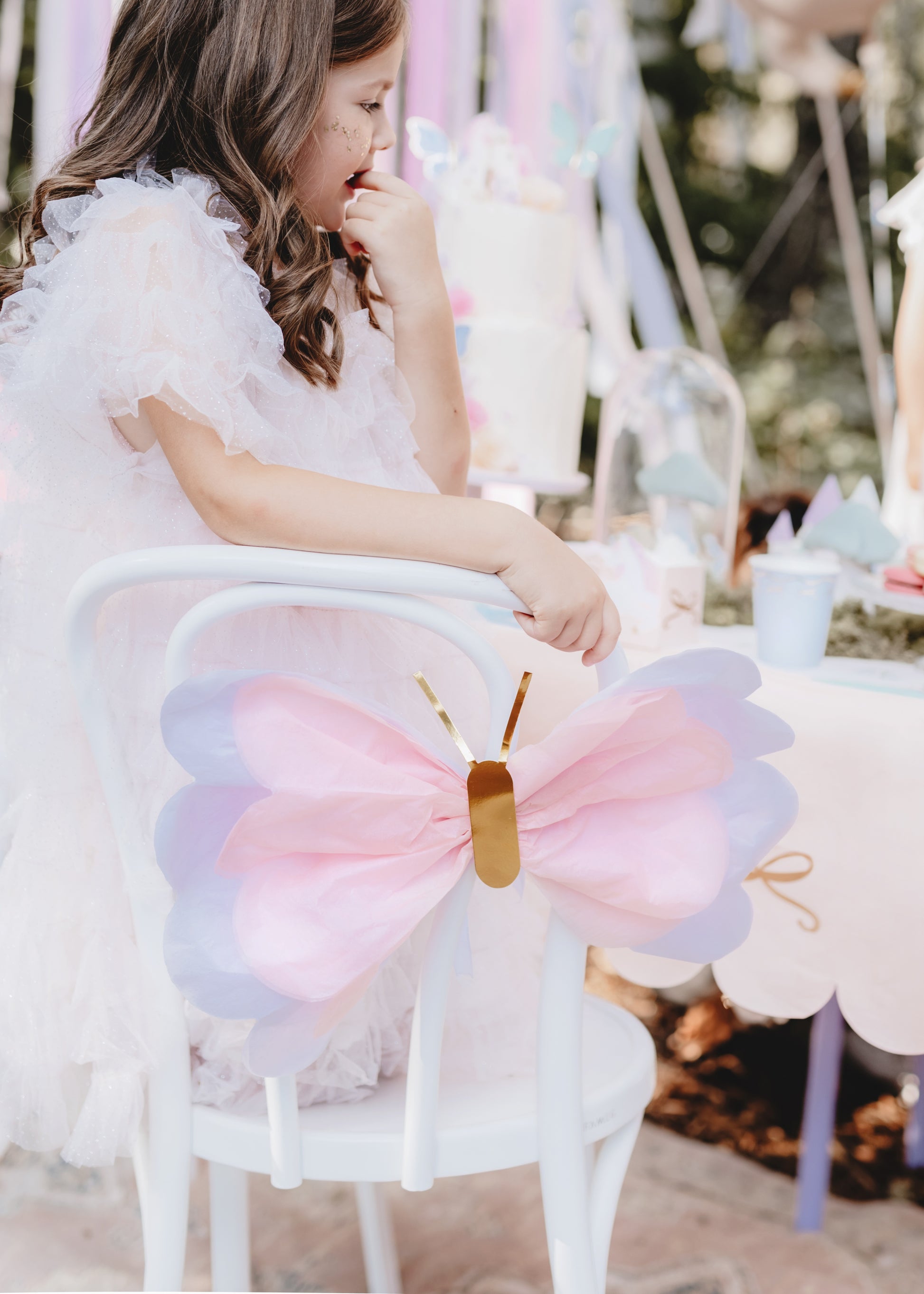 Child in a white dress with a large bow sitting on a chair at a party.