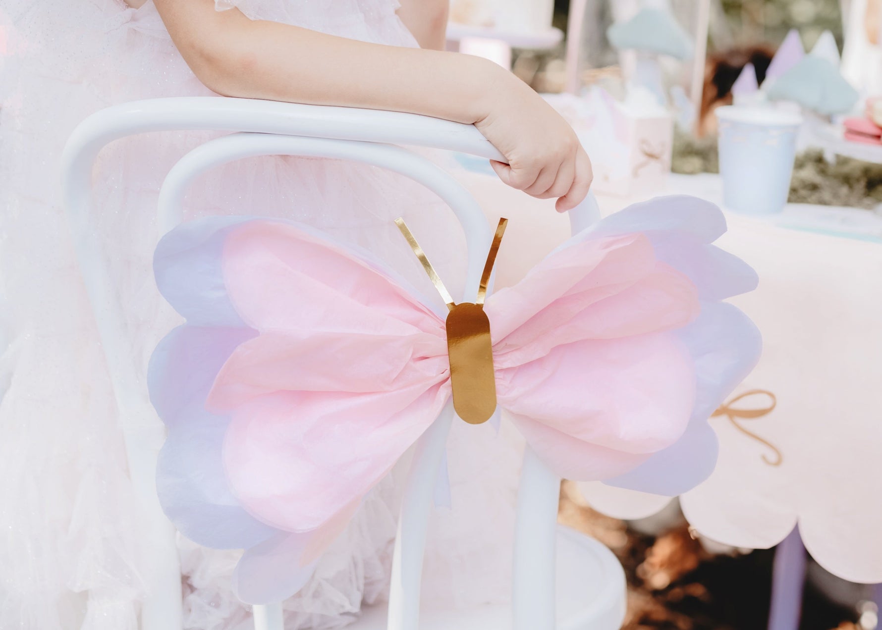 Child in a white dress with a large bow sitting on a chair at a festive outdoor event.