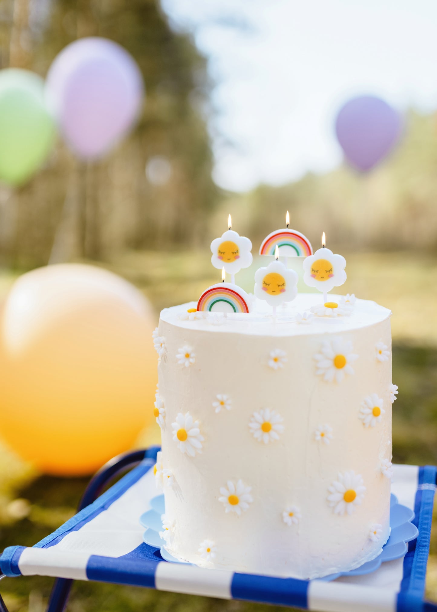 Small white cake with colorful daisy and rainbow candles on a blue and white striped stand, with balloons in the background.