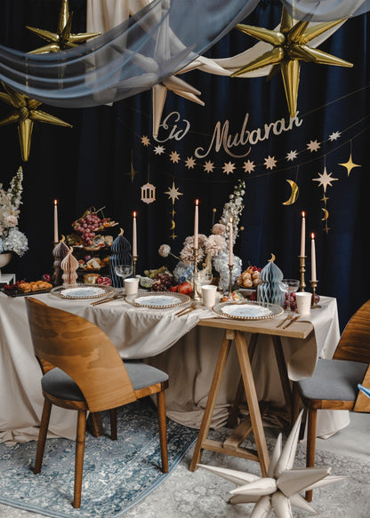 Decorated dining table with 'Eid Mubarak' text, gold stars, and festive decorations against a dark wall.