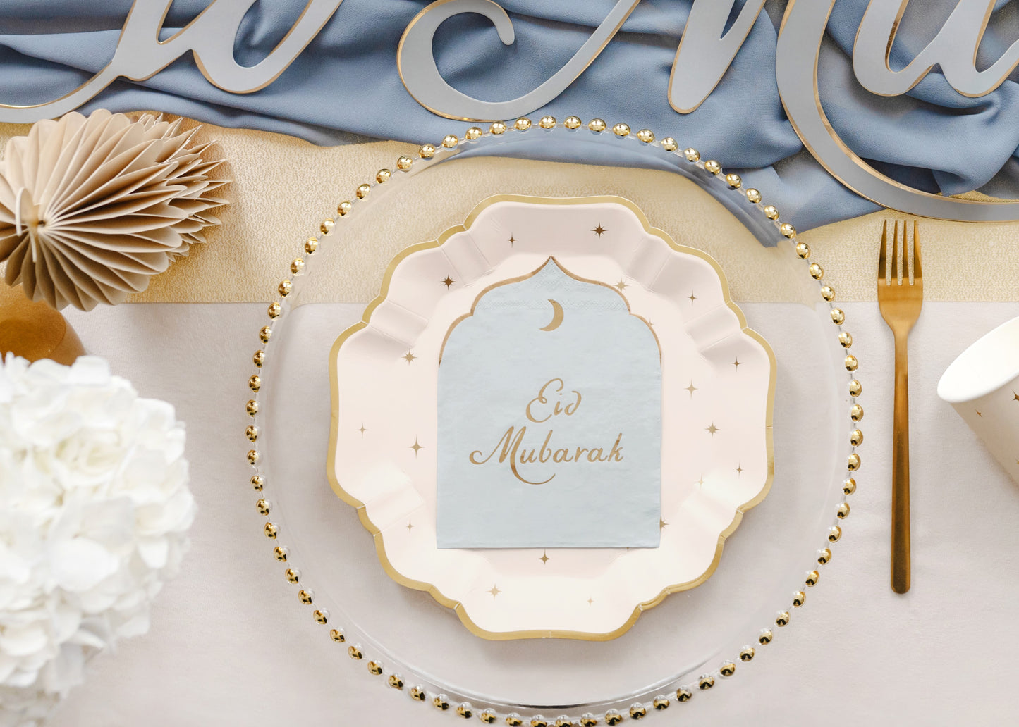 Decorative table setting with 'Eid Mubarak' napkin on a plate, surrounded by flowers and gold cutlery.