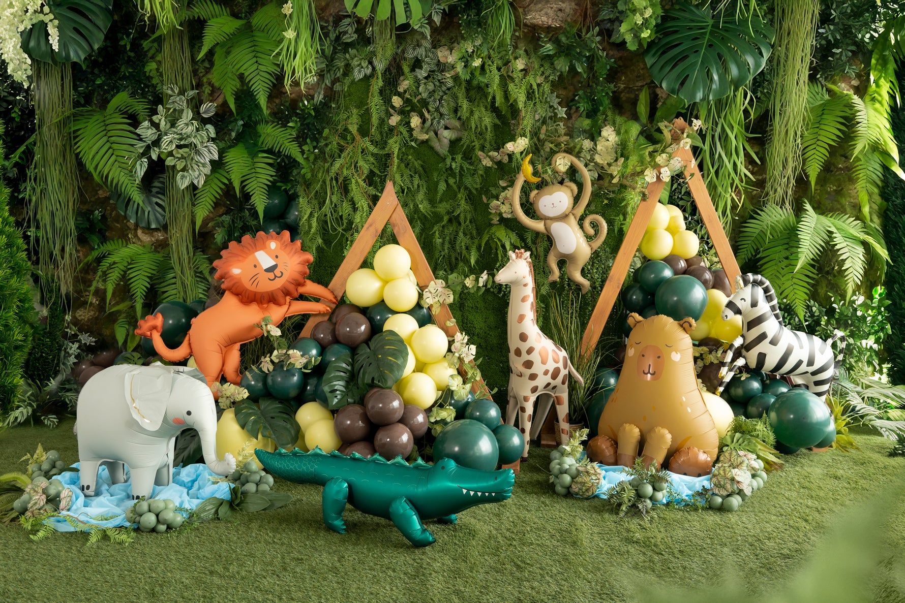Foil balloons in shape of animals including a lion, elephant, giraffe, and zebra in a jungle setting with plants and grass.