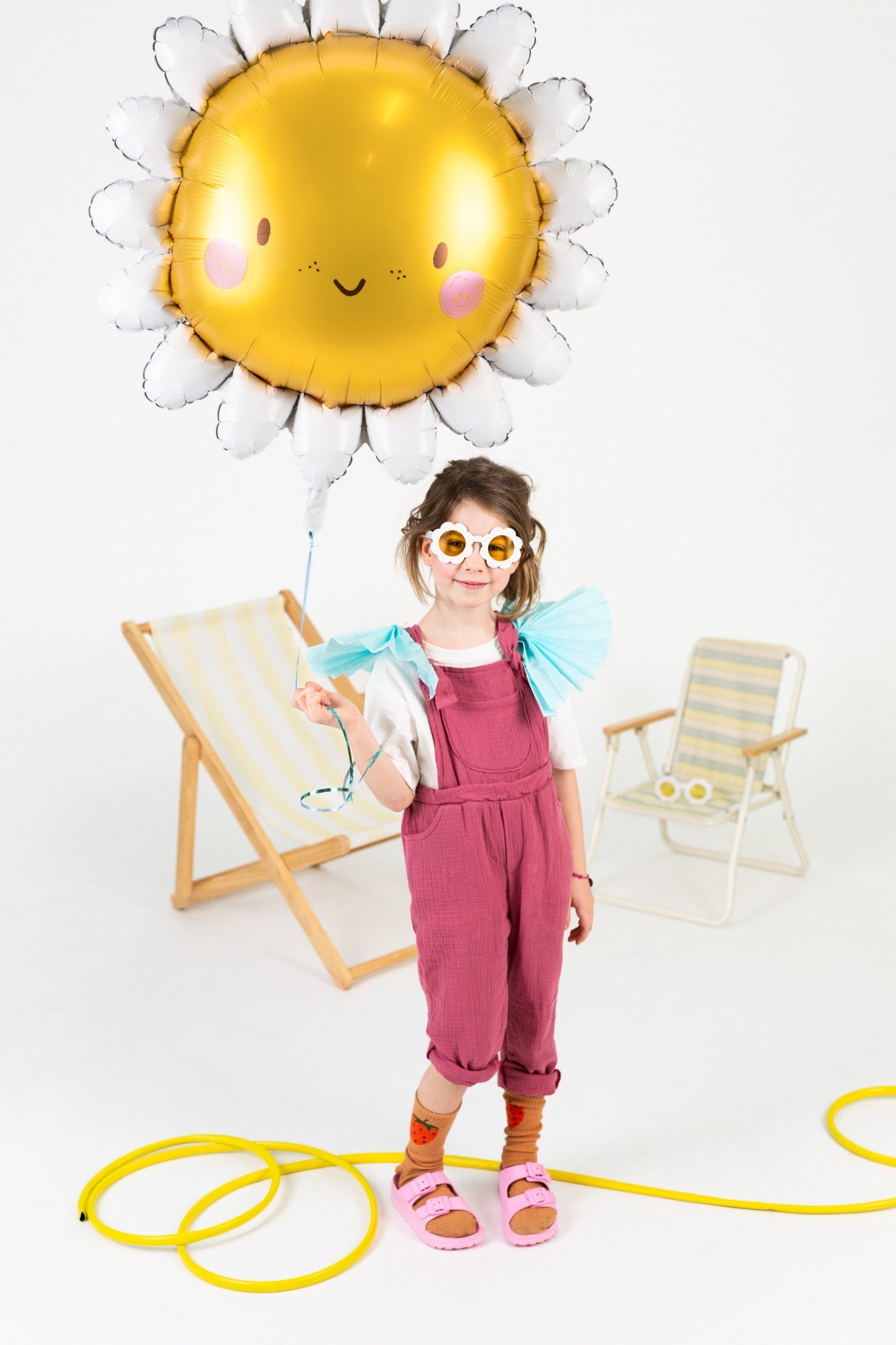 Girl holding a big shaped sun foil balloon, wearing flower sunglasses, in a bright summer setup