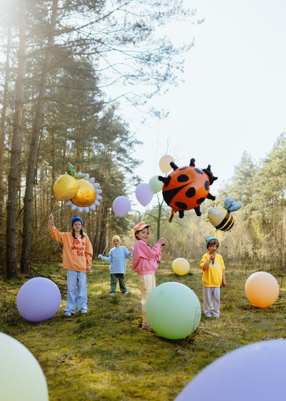 Kids playing outdoors with large foil balloons in shapes of sun, ladybug and bee, forest background