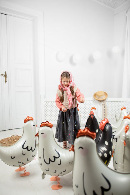 Little girl in traditional village costume playing with walking chicken and hen foil balloons indoors, surrounded by white and black balloons shaped like hens