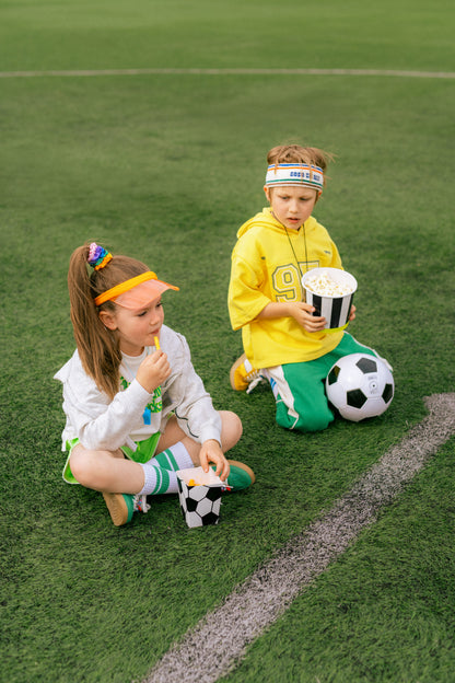 Two children sitting on a soccer field with sports gear and popcorn.