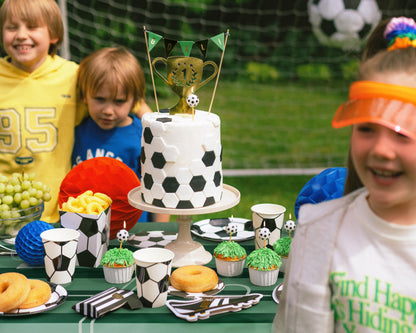 Children around a table with a soccer-themed cake and snacks outdoors.