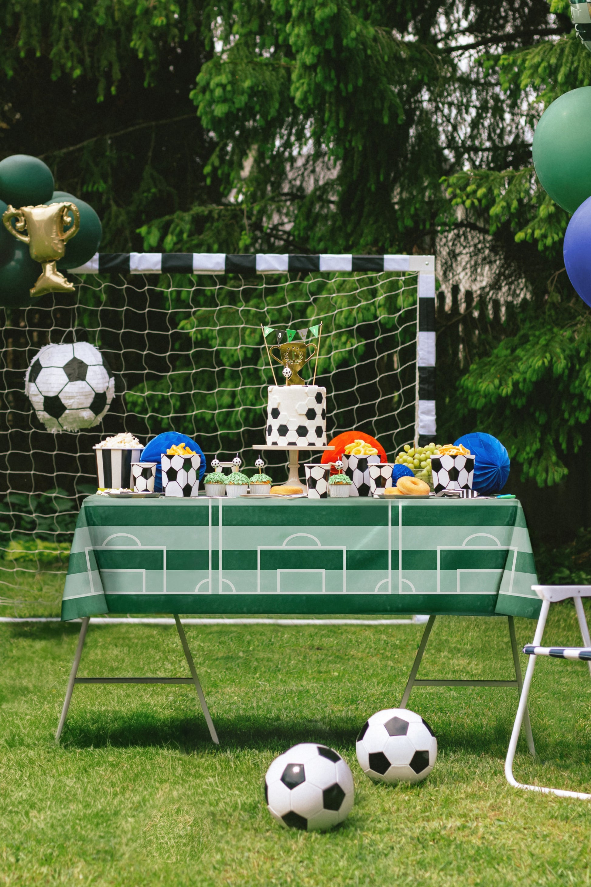 Children's party setup with soccer-themed decorations, including a table with small soccer balls and trophies, and a goal in the background.