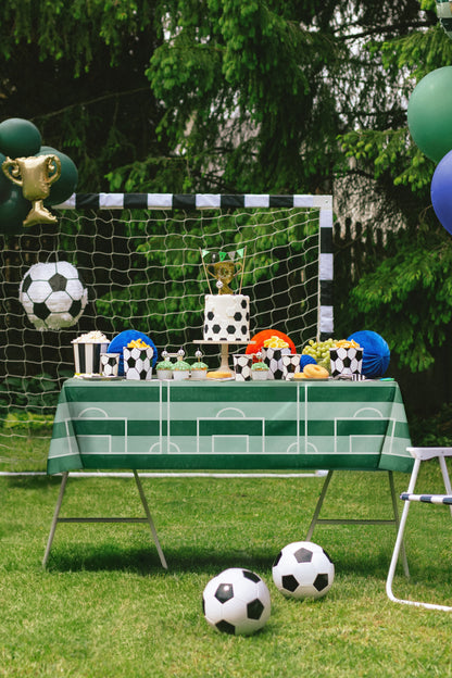 Children's party setup with soccer-themed decorations, including a table with small soccer balls and trophies, and a goal in the background.