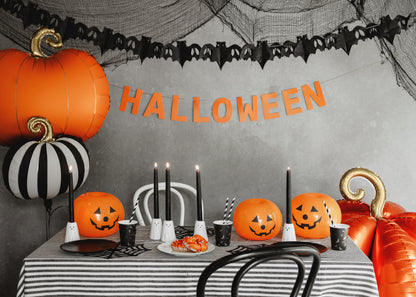 Halloween-themed table setting with pumpkins, candles, and decorations against a gray wall with 'HALLOWEEN' banner.