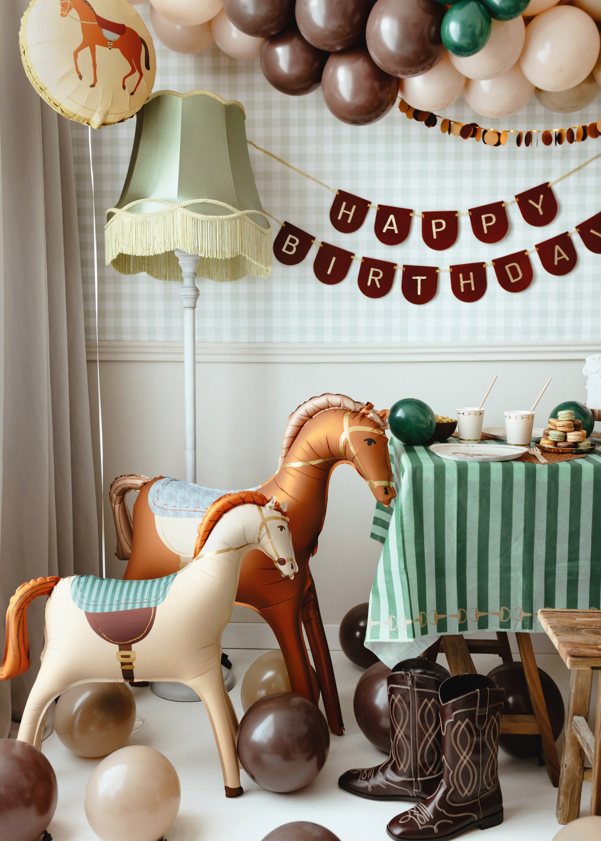 Western-themed kids' birthday party setup with inflatable horse balloons, brown and beige latex balloons, cowboy boots, and green striped tablecloth