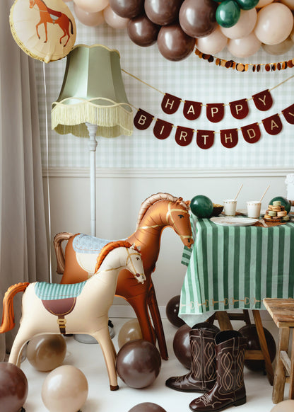 Western-themed kids' birthday party setup with inflatable horse balloons, brown and beige latex balloons, cowboy boots, and green striped tablecloth