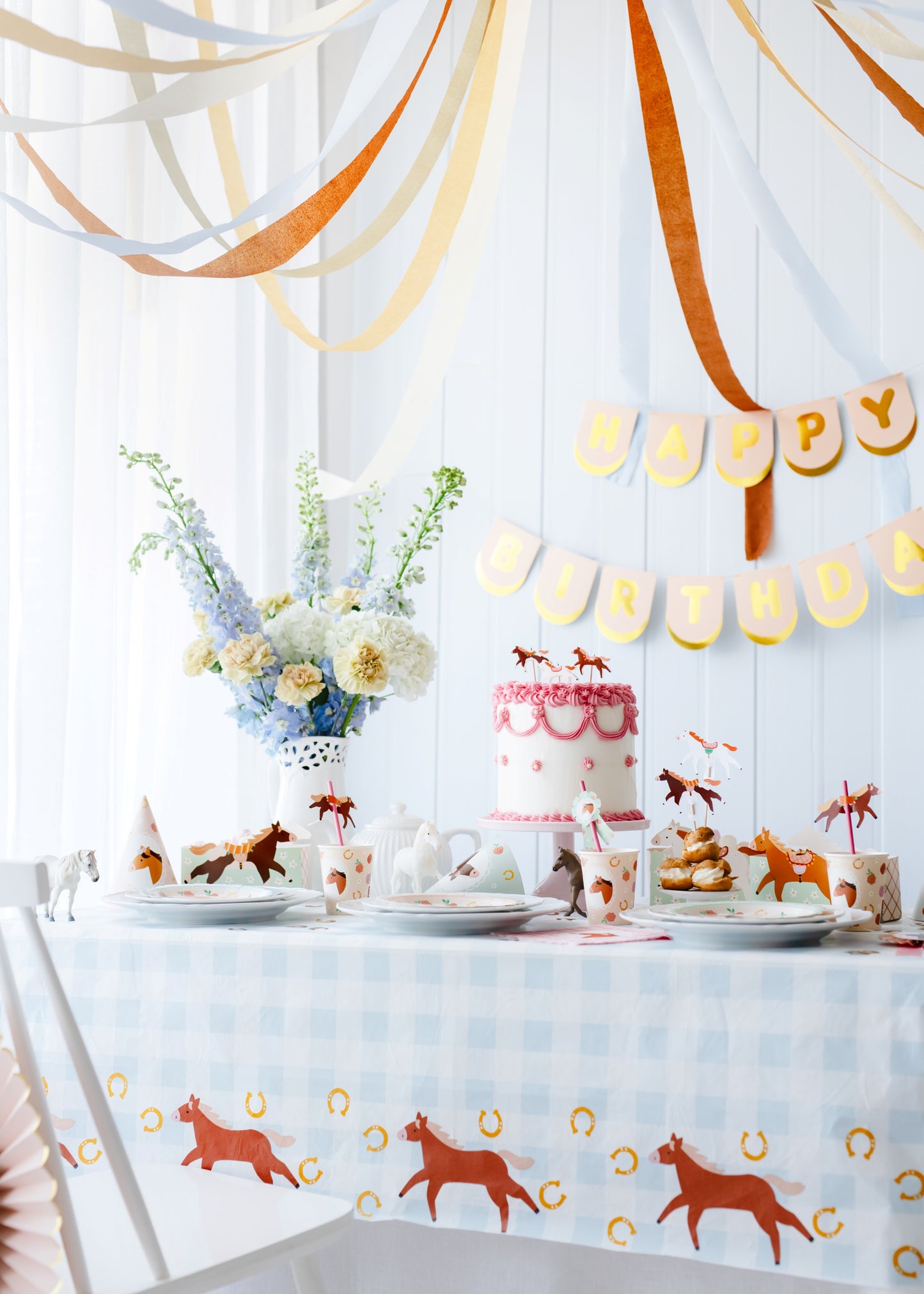 Decorated table with cake, plates, and animal-themed decorations against a white wall with hanging ribbons and 'Happy Birthday' banner.