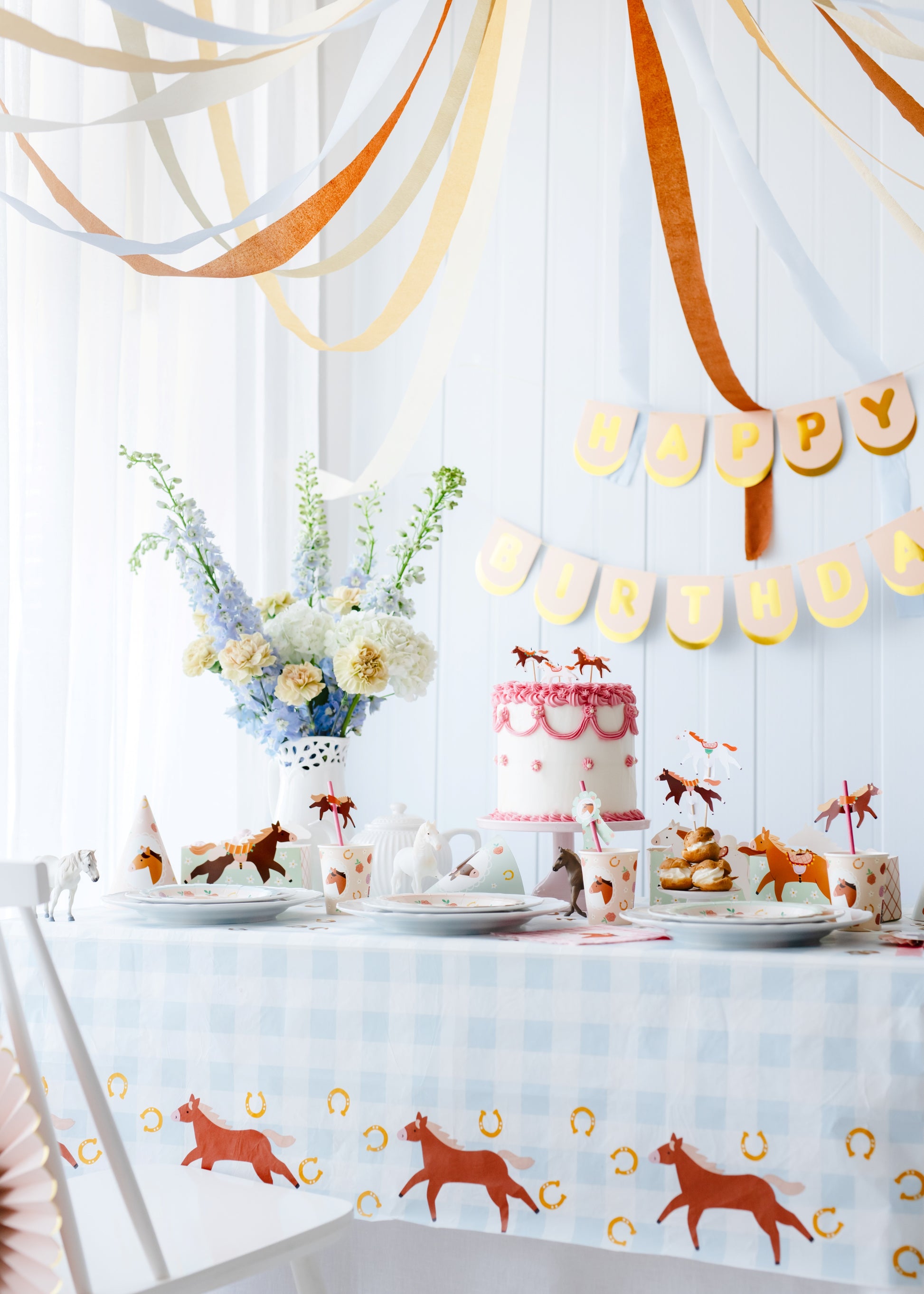 Decorated table with cake, plates, and animal-themed decorations against a white wall with hanging ribbons and 'Happy Birthday' banner.