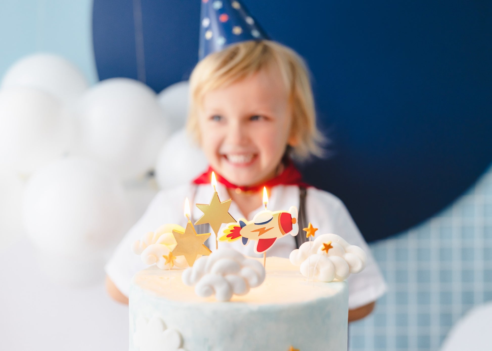 Rocket and star candles on a birthday cake with a smiling child wearing a party hat in the background