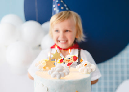 Rocket and star candles on a birthday cake with a smiling child wearing a party hat in the background