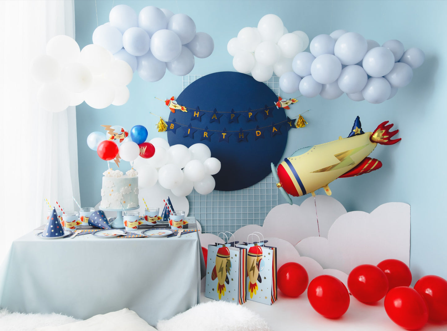 Complete rocket-themed birthday setup with cake, red balloons, foil rocket balloon, and decorated table surrounded by clouds
