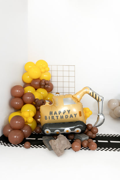 Foil balloon shaped like an excavator with 'Happy Birthday' text, surrounded by yellow and brown balloon decorations