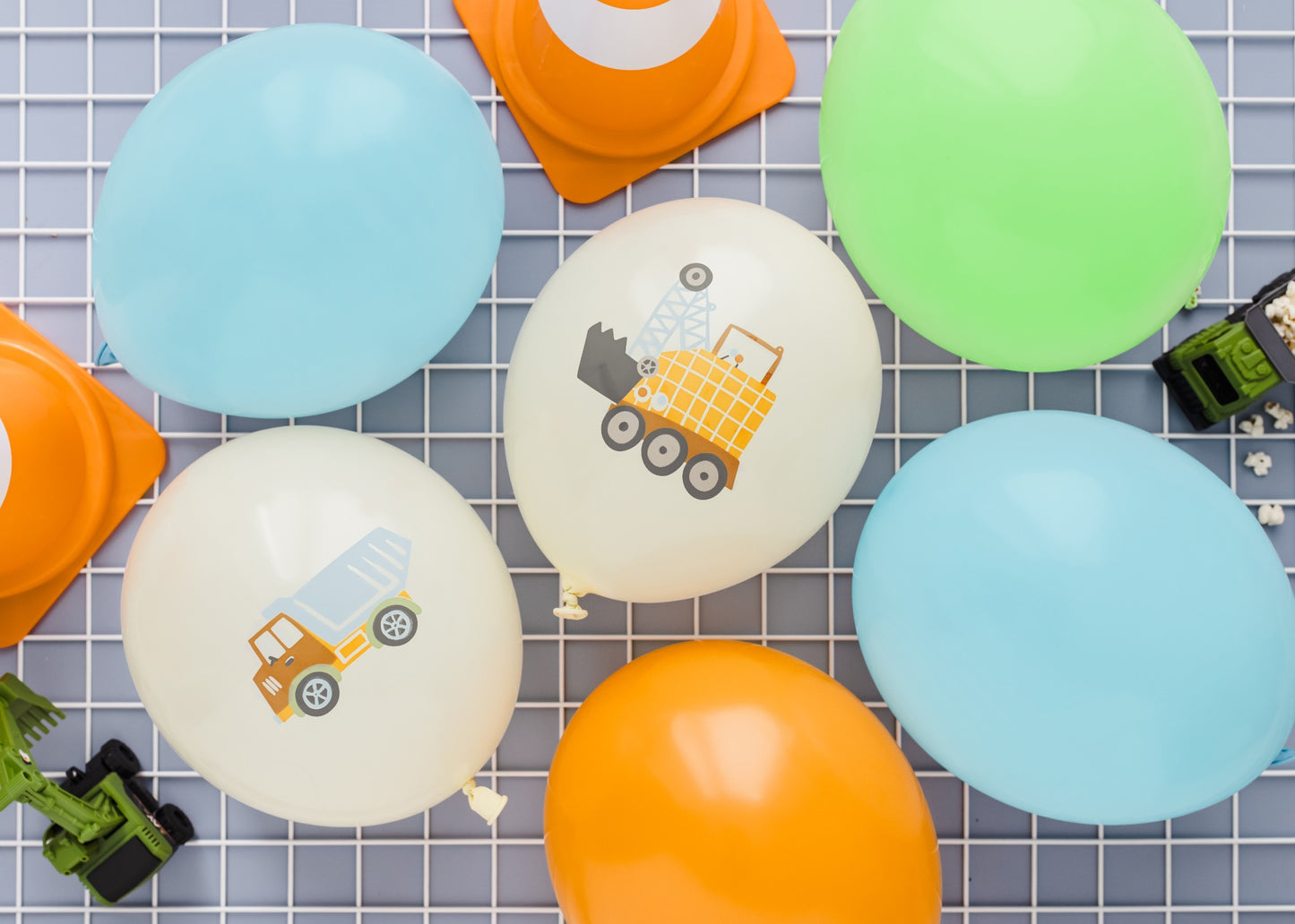 Colorful latex balloons decorated with construction vehicle illustrations, displayed with orange traffic cones and toy trucks