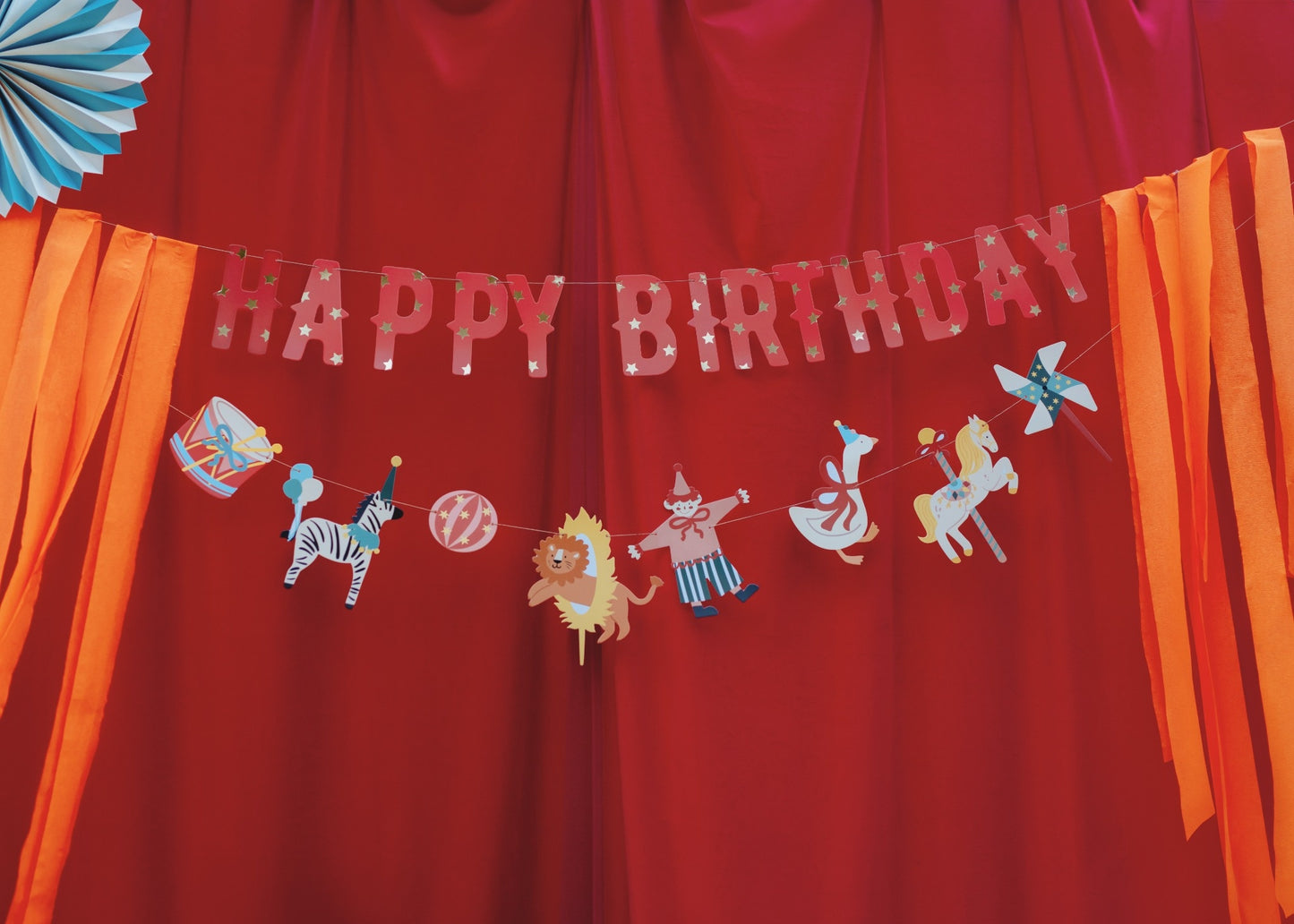 Colorful circus-themed 'Happy Birthday' banner with illustrations of animals, circus elements and hanging orange ribbons on both sides