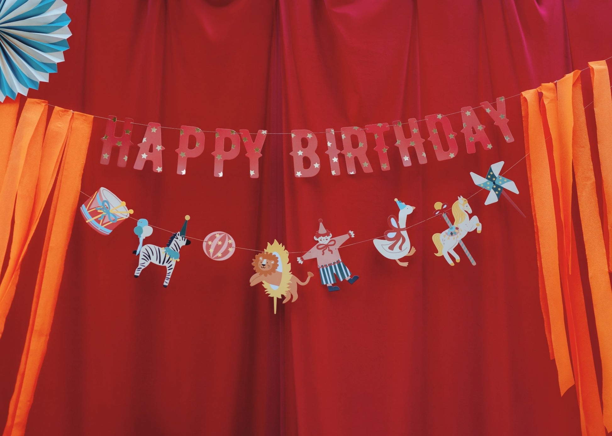 Colorful circus-themed 'Happy Birthday' banner with illustrations of animals, circus elements and hanging orange ribbons on both sides