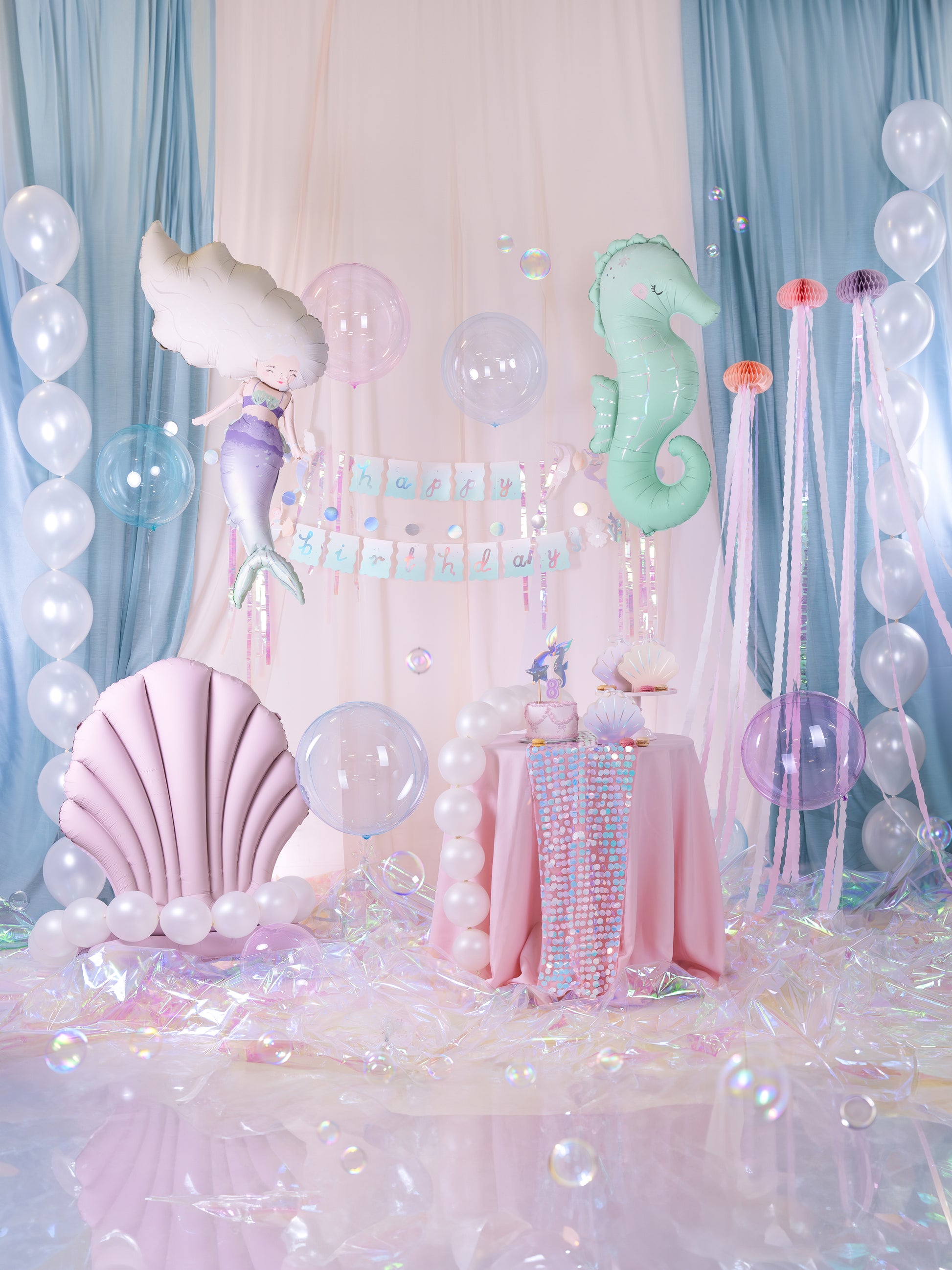 Under the sea mermaid birthday party setup with pastel mermaid and seahorse foil balloons, pink seashell balloon, iridescent decorations, jellyfish hanging streamers, balloon arch, 'Happy Birthday' banner and a pink table with themed cake and seashell plates.