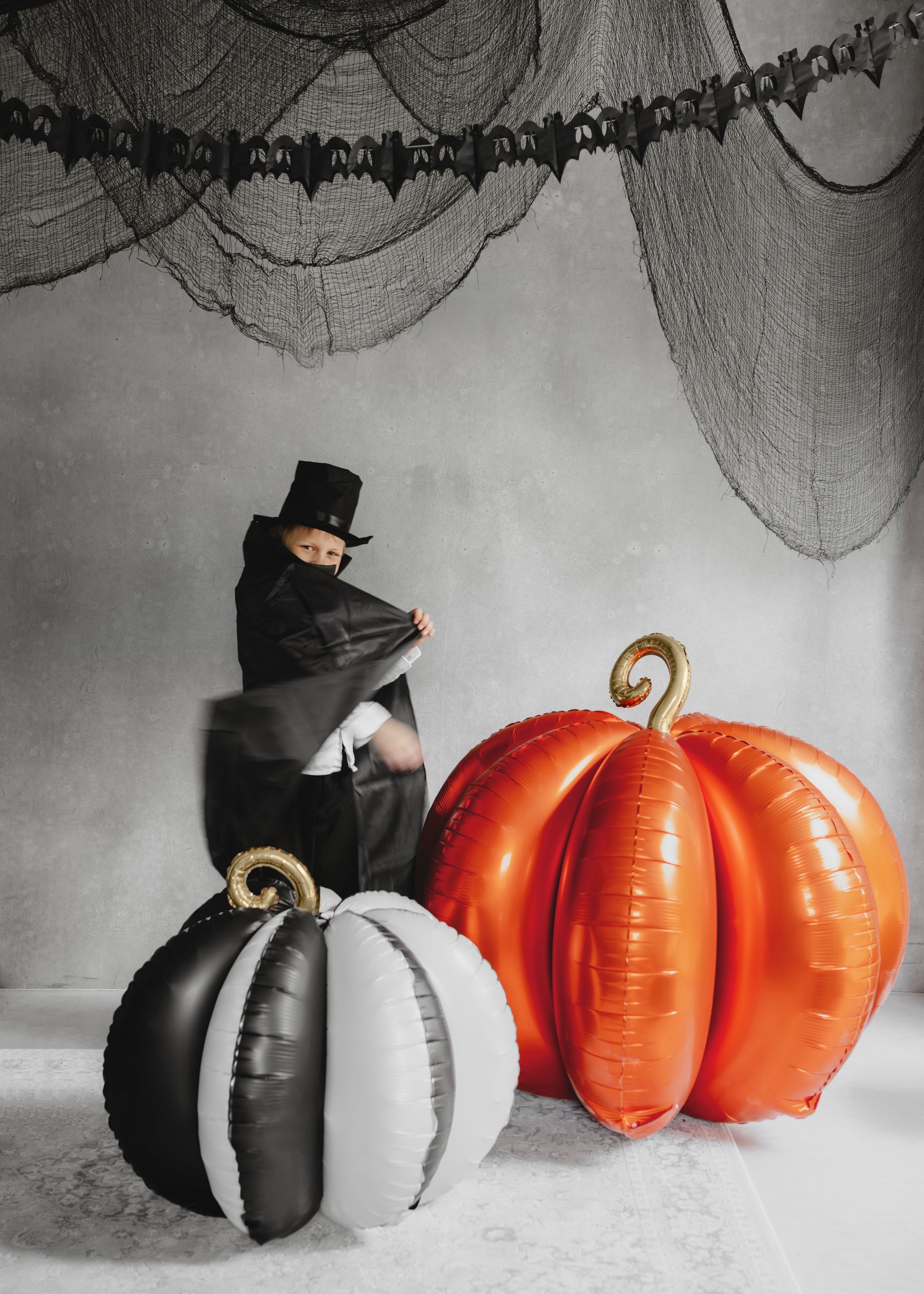 Person in a black outfit with a top hat next to inflatable pumpkins on a Halloween-themed background.