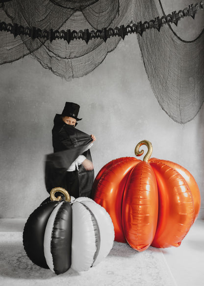 Person in a black outfit with a top hat next to inflatable pumpkins on a Halloween-themed background.