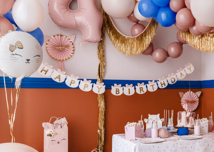 Cat-themed birthday party decorations with a "Happy Birthday" banner, pastel and metallic balloons, gold garland, cat face gift bags, snack boxes, and pink cake on a festive table, perfect for a kids' kitty party.