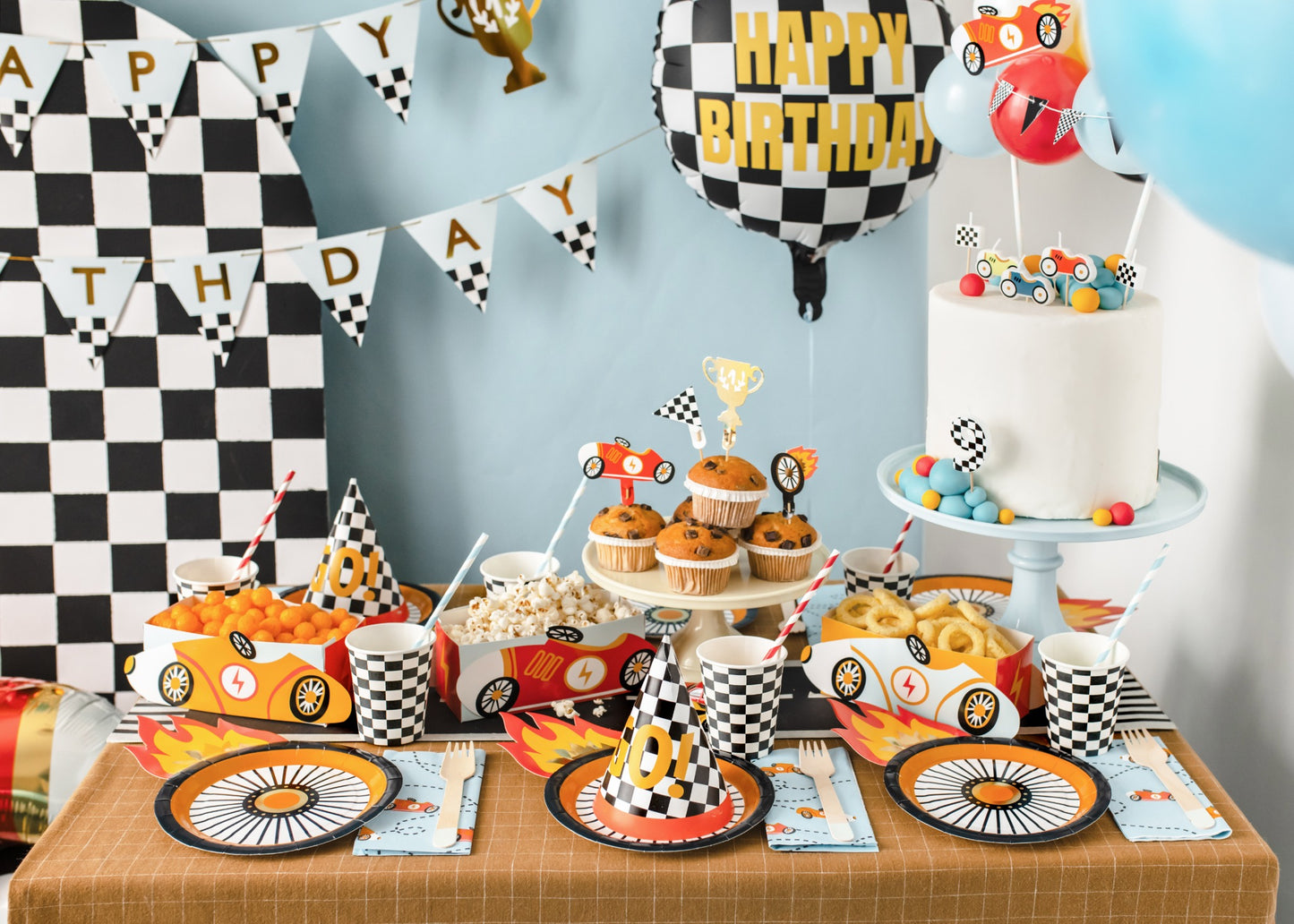 Racing car themed party table with checkered paper cups, wheel-shaped plates, popcorn snack boxes shaped like race cars, muffins, and colorful flame decorations.
