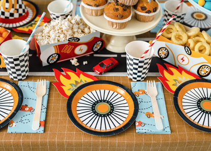 Racing car themed party table with checkered paper cups, wheel-shaped plates, popcorn snack boxes shaped like race cars, muffins, and colorful flame decorations.