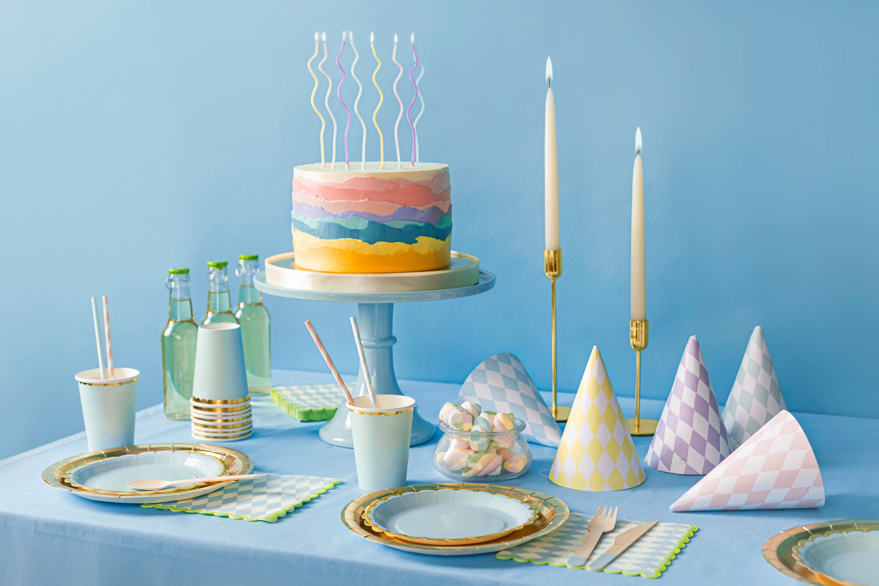 Pastel-colored birthday cake with candles on a table with paper cups and plates.