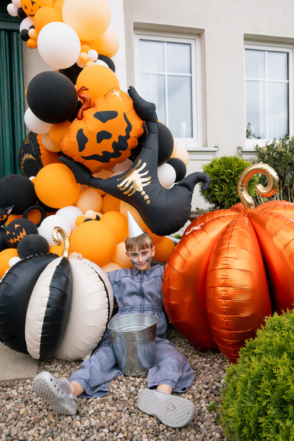A child in a gray jumpsuit and party hat sits on a gravel surface in front of a house, surrounded by large orange, black, and white Halloween balloon decorations, including pumpkins, a ghost, and a metallic bucket.