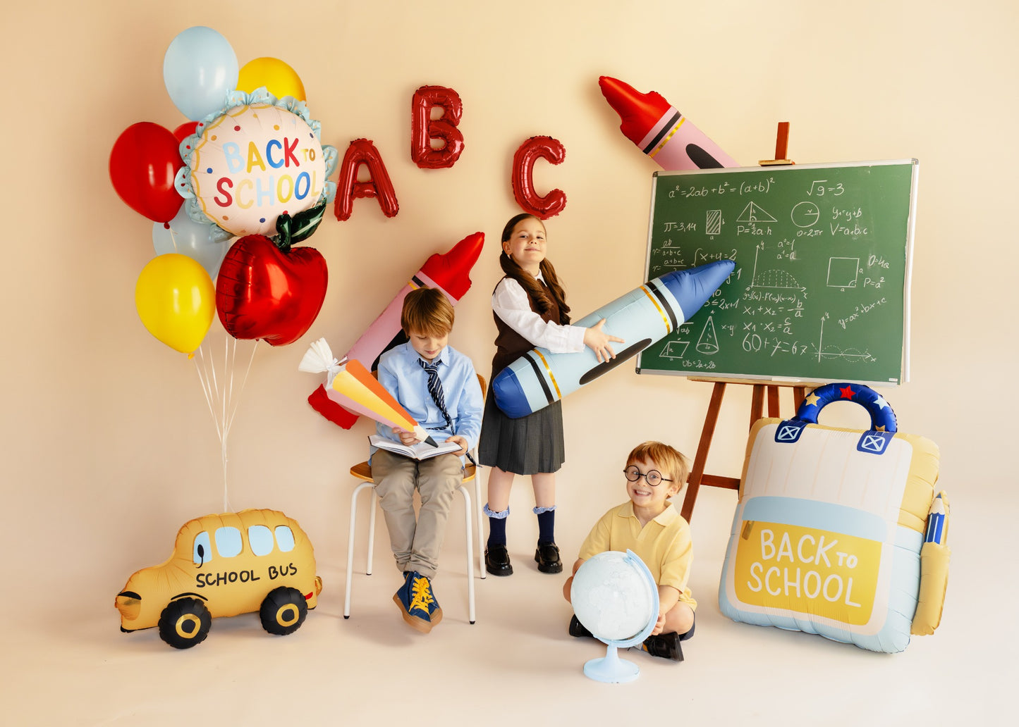 A school-themed party scene with children playing with educational props including a chalkboard, balloons, a toy school bus, and a large balloon backpack labeled BACK to SCHOOL, set against a warm beige wall.