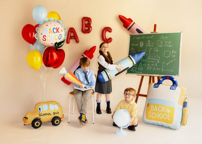 A school-themed party scene with children playing with educational props including a chalkboard, balloons, a toy school bus, and a large balloon backpack labeled BACK to SCHOOL, set against a warm beige wall.
