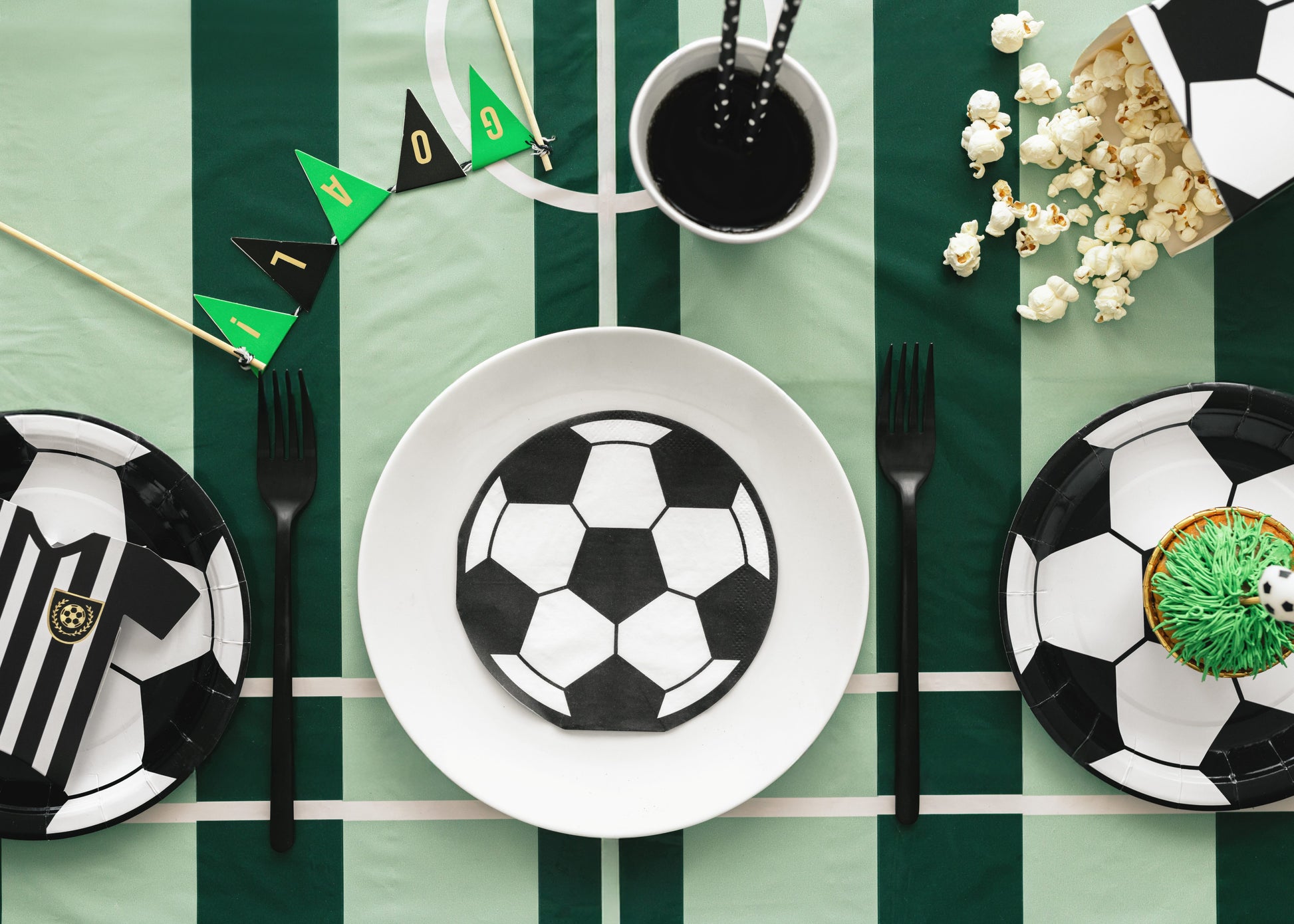 Soccer-themed table setting with plates, cutlery, and snacks on a green and white striped tablecloth.