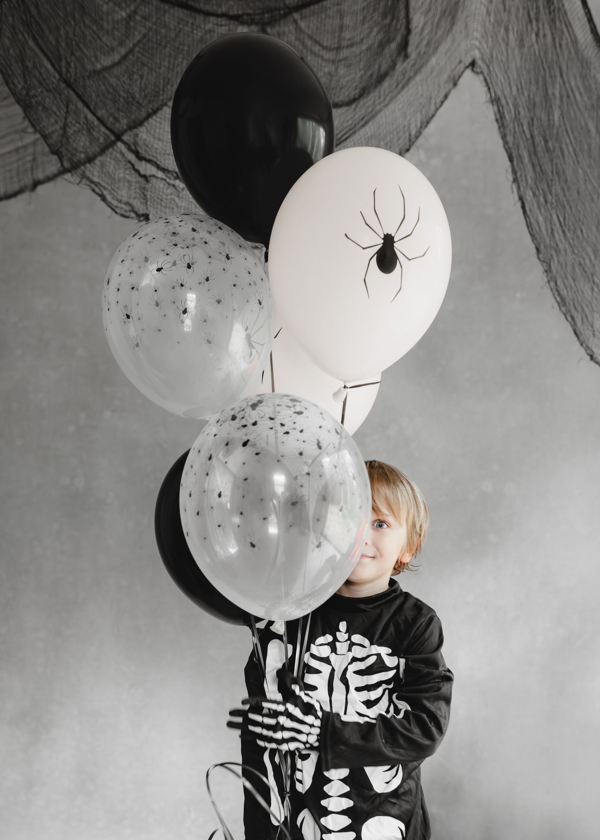 Child in a skeleton outfit holding Halloween-themed balloons with black and white colors.