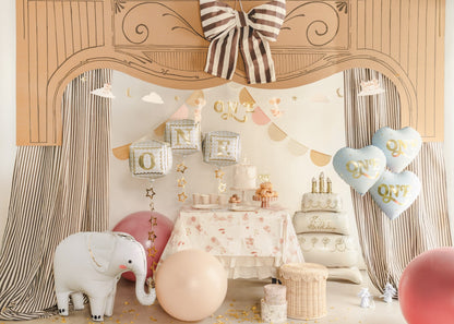 Decorative setup with balloons, cake, and elephant toy in a room with curtains and a decorative arch.