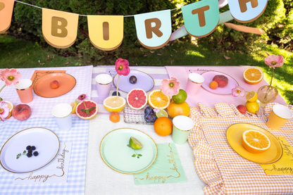 Colorful outdoor birthday table setup with gingham tablecloths, pastel party plates, cups, citrus fruits, and floral decor – summer picnic party tableware