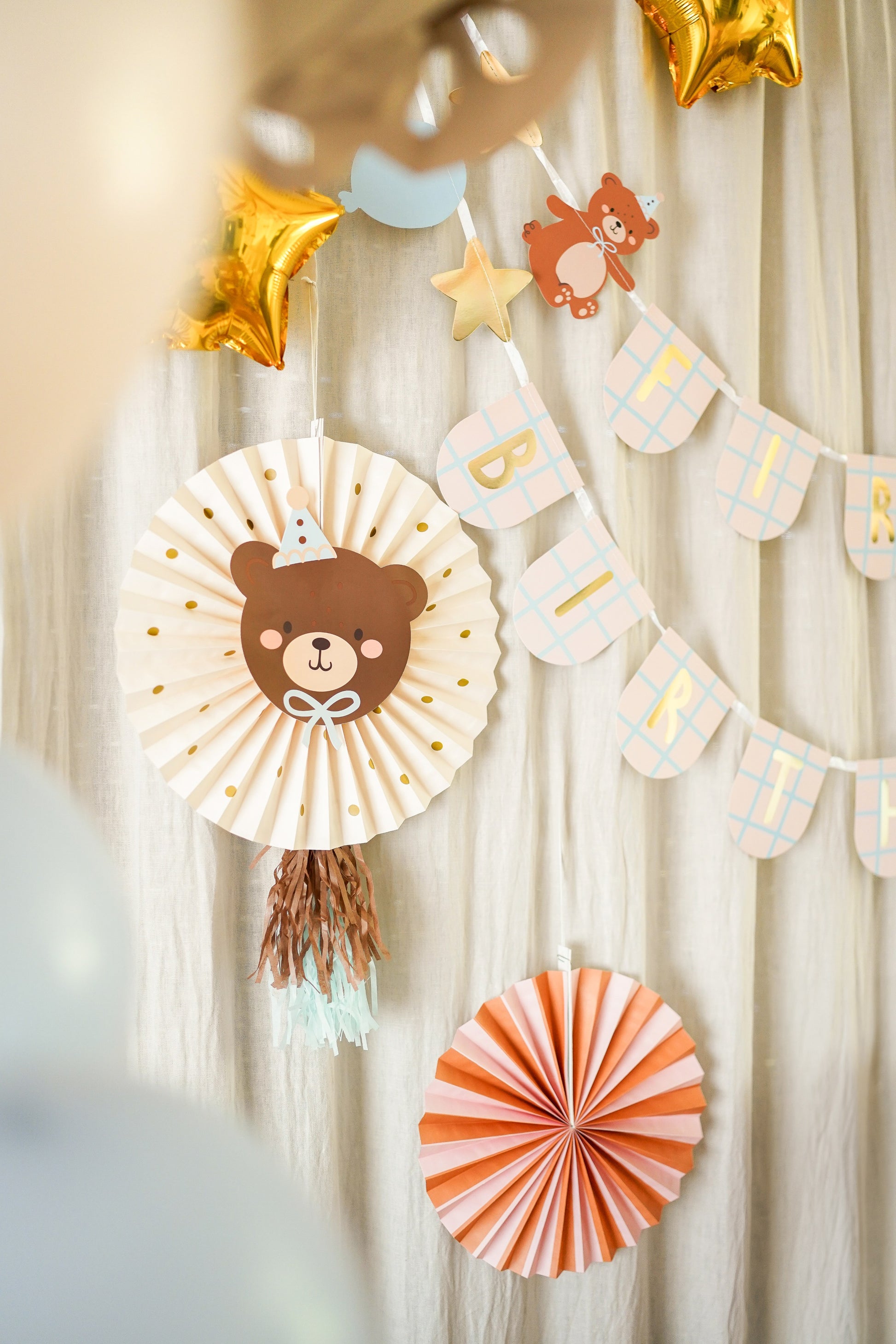 Decorative wall with bear-themed paper fan, star-shaped balloons, and banner against a neutral curtain background.