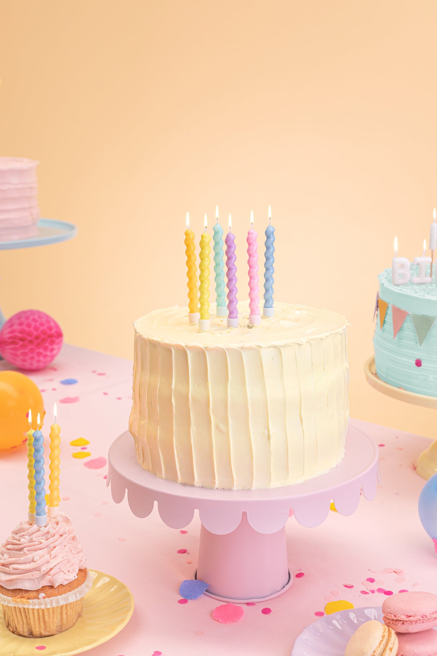 Pastel-colored birthday cake with candles on a pink stand against a yellow background
